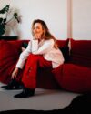 Leonie Young sitzt auf einem roten Sofa, trägt eine weiße Bluse und rote Hose und lächelt.